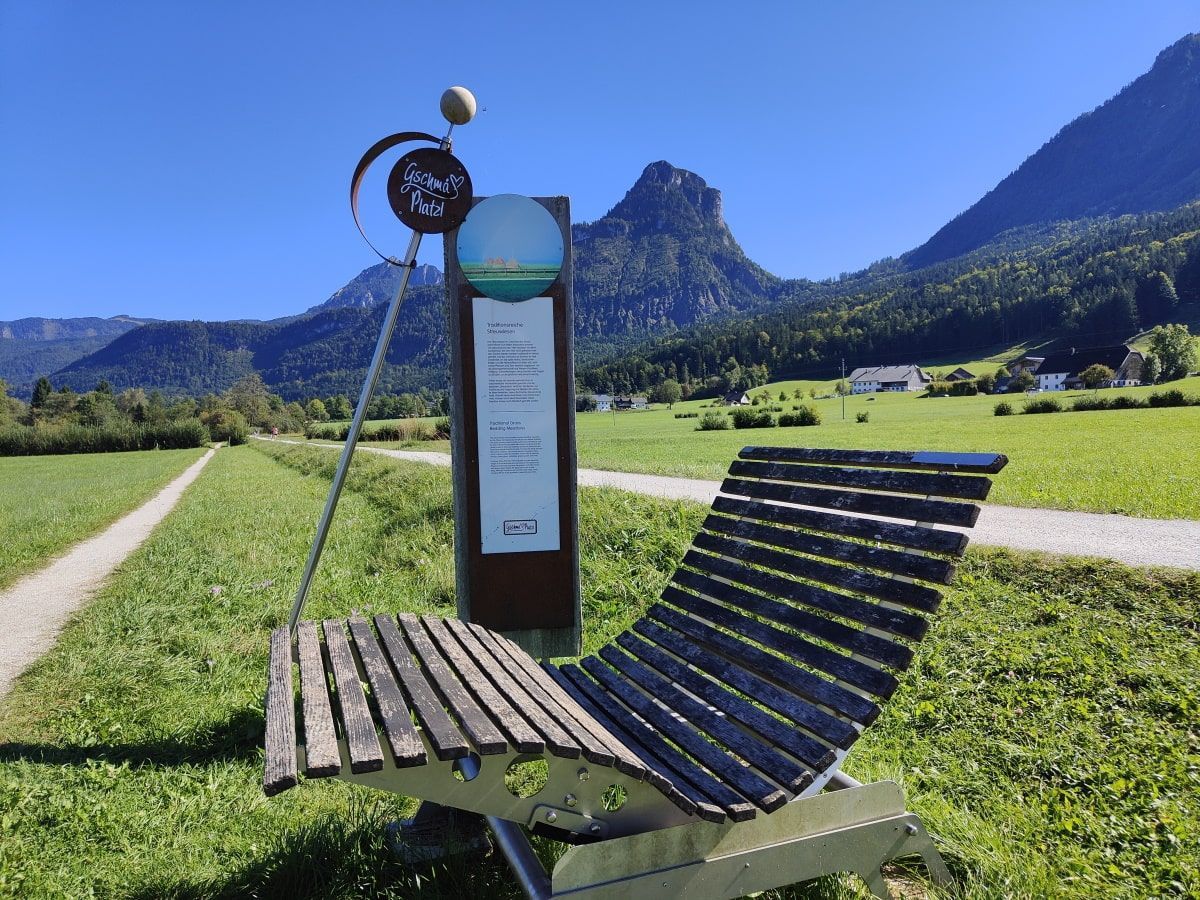 Gschma Platzl Salzkammergut Wooden lounge chair by a sign with a mountain backdrop on a sunny day.