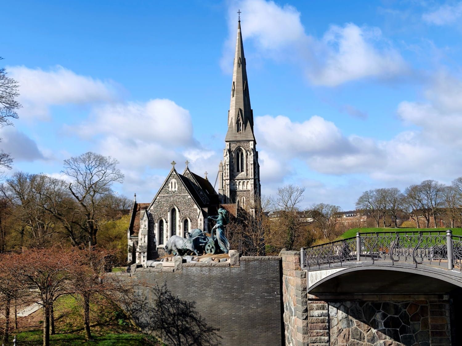 Sankt Albans Kirche Stone church with tall spire beside a bridge, under a blue sky with clouds and trees.