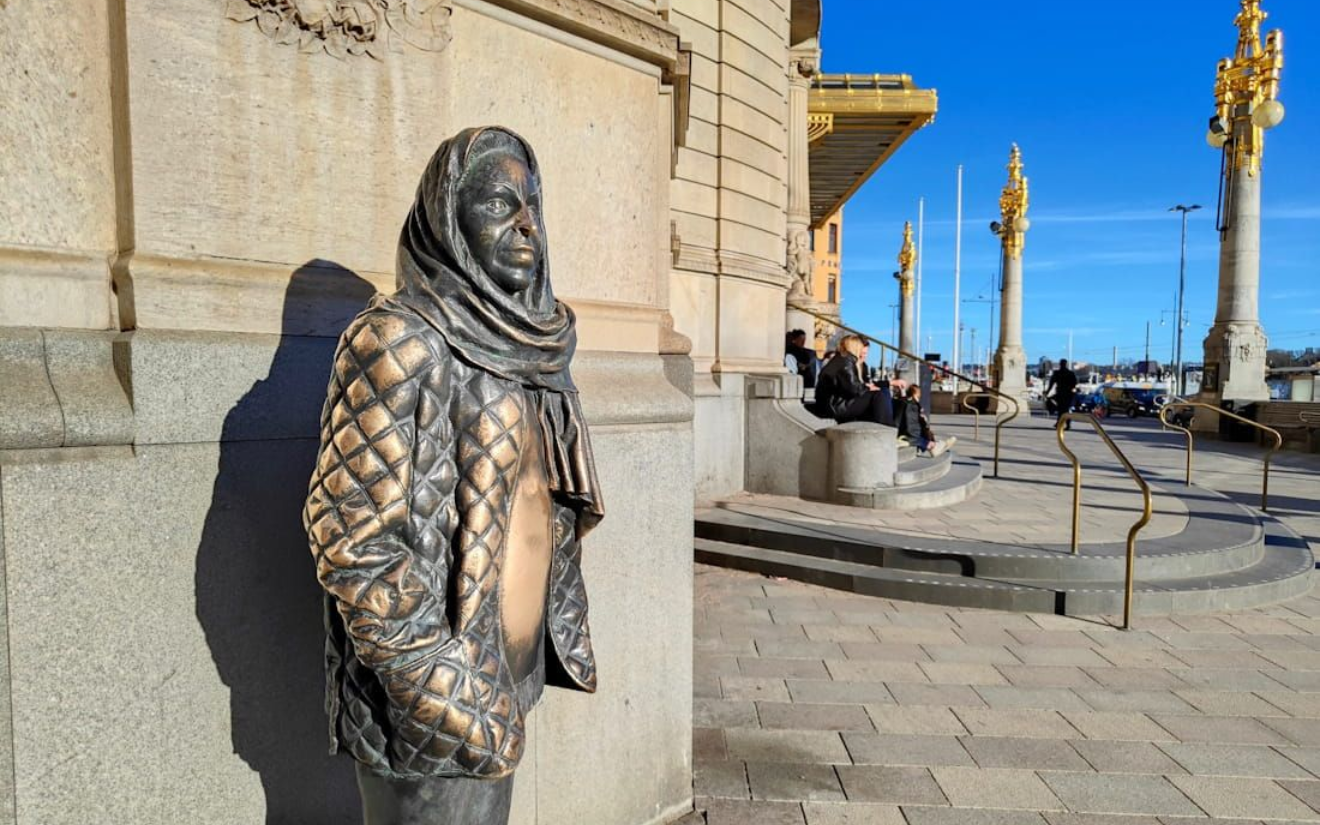 Schauspielerin Margareta Krook Statue Bronze hooded statue by a stone wall on a sunny city promenade with ornate lamp posts. Schauspielerin Margareta Krook Statue.
