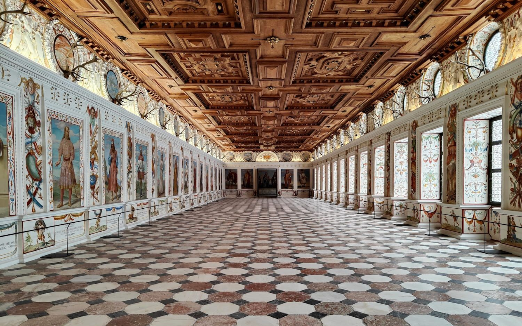 Schloss Ambras Innsbruck Long, ornate hall with patterned floor, decorated walls, and carved wooden ceiling.