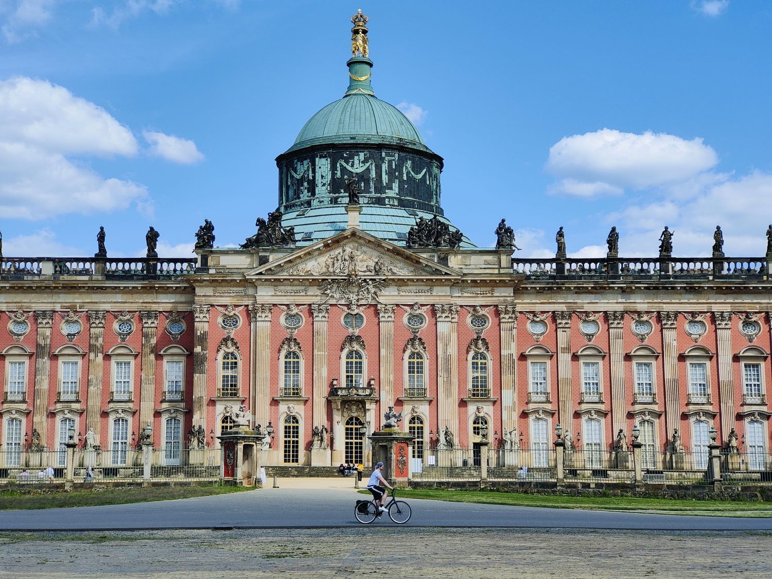 Neues Palais Potsdam Palace facade, reddish-brown with a turquoise dome, set against a blue sky. People on bikes in front.