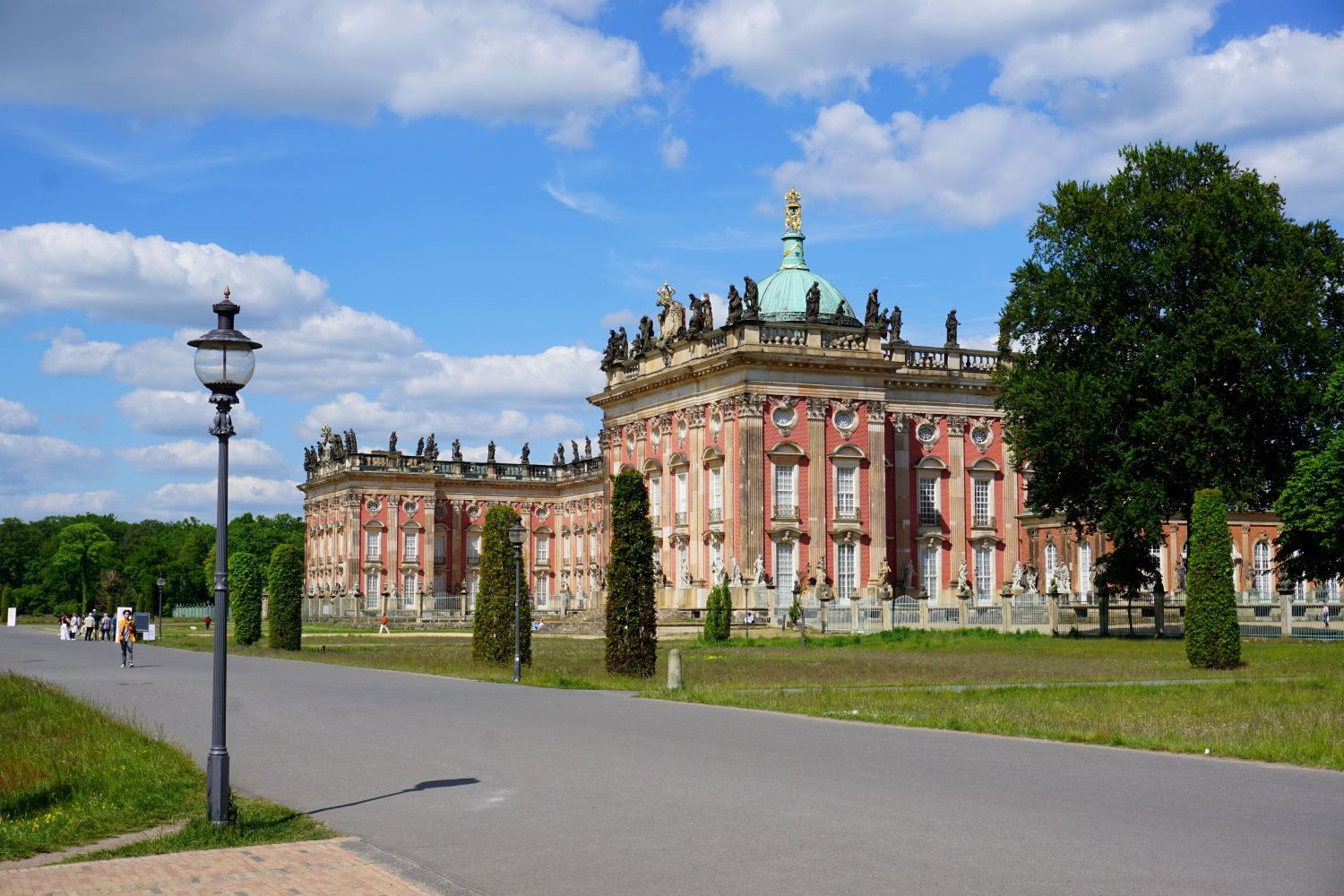 Ornate reddish-brown and white palace with green dome under blue sky, road and grass in foreground.