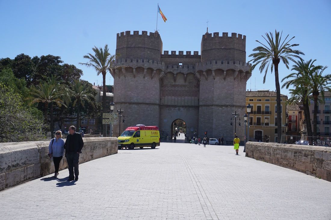 View of Serranos Gate in Valencia, Spain. Two people walk on a stone bridge toward the medieval structure. An ambulance is present.