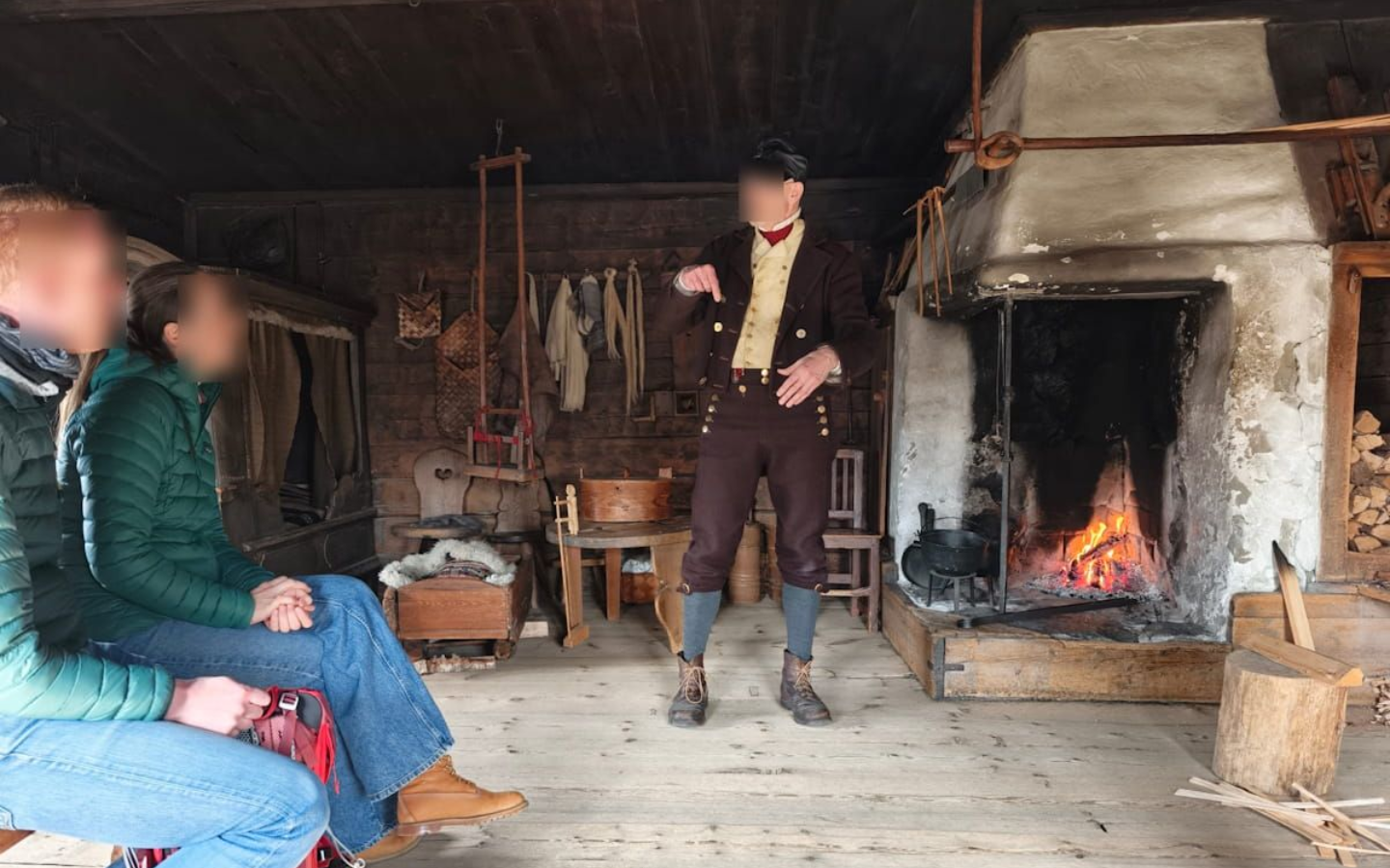 Skansen Stockholm Two people sitting by a fireplace in a rustic room, with a third person standing near the hearth. Skansen Stockholm.