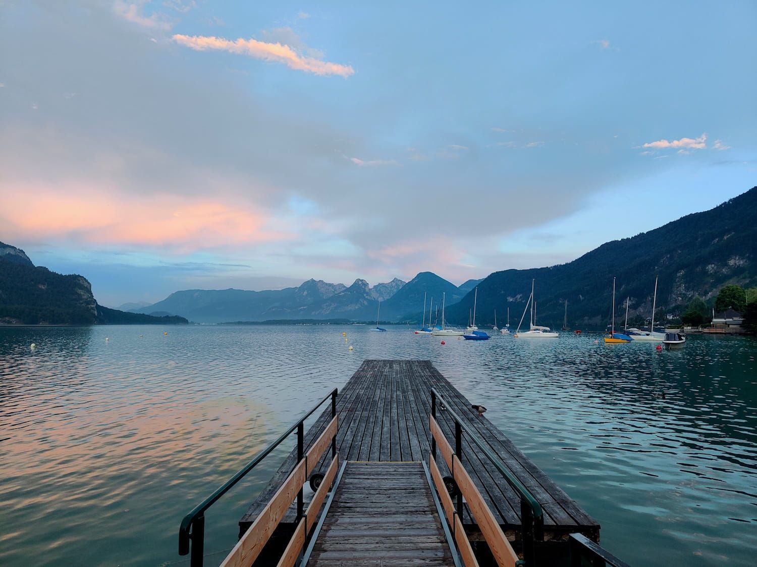 Ein hölzerner Steg erstreckt sich bei Sonnenuntergang in einen ruhigen See, in der Ferne sind Berge und Segelboote zu sehen.