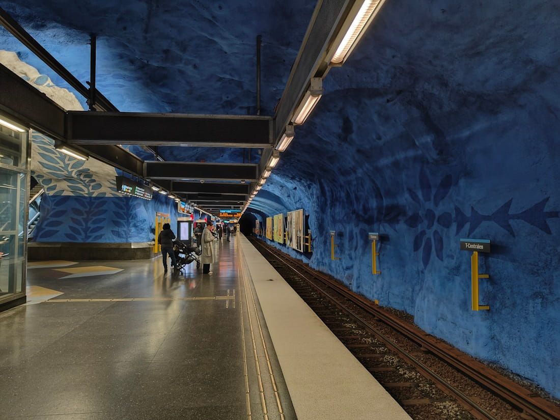 Underground subway platform with blue rock walls, train tracks, and a few people walking