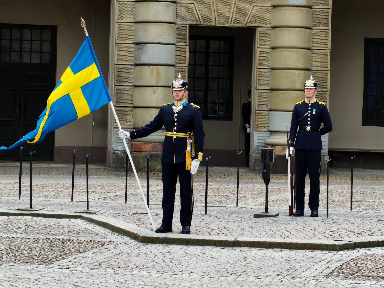 Two guards in dark uniforms stand at attention beside a large Swedish flag outside a stone building