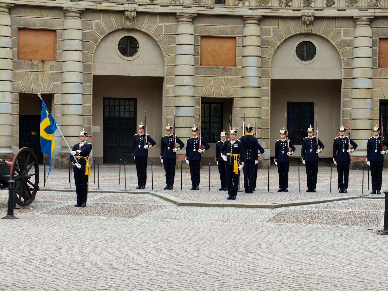 Military honor guard lined up before a historic stone building, with a blue-yellow flag at left.