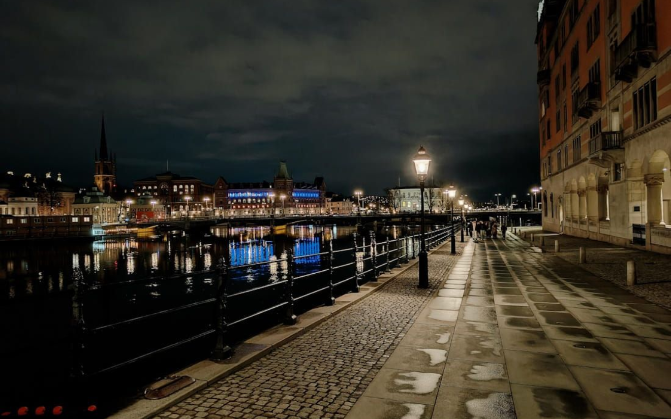 Rathaus Stockholm Nighttime riverside promenade with wet cobblestones, lit street lamps, and city lights reflecting on the water.