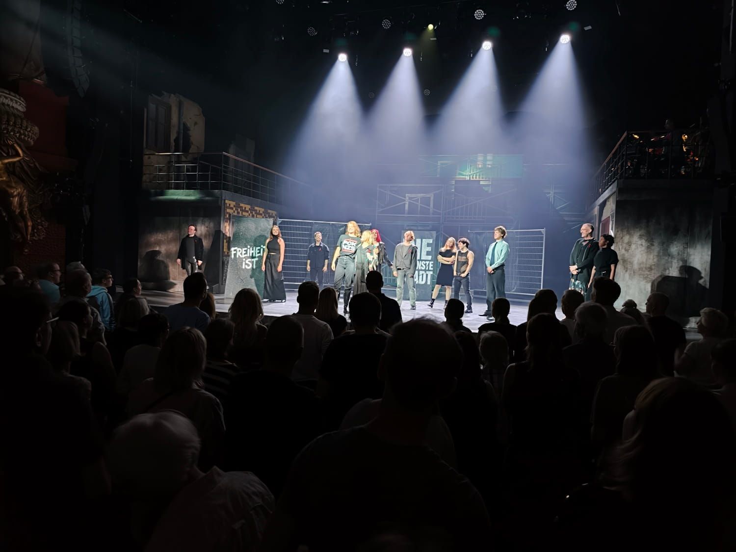 Performers on a stage under bright spotlights, with a dark audience in the foreground