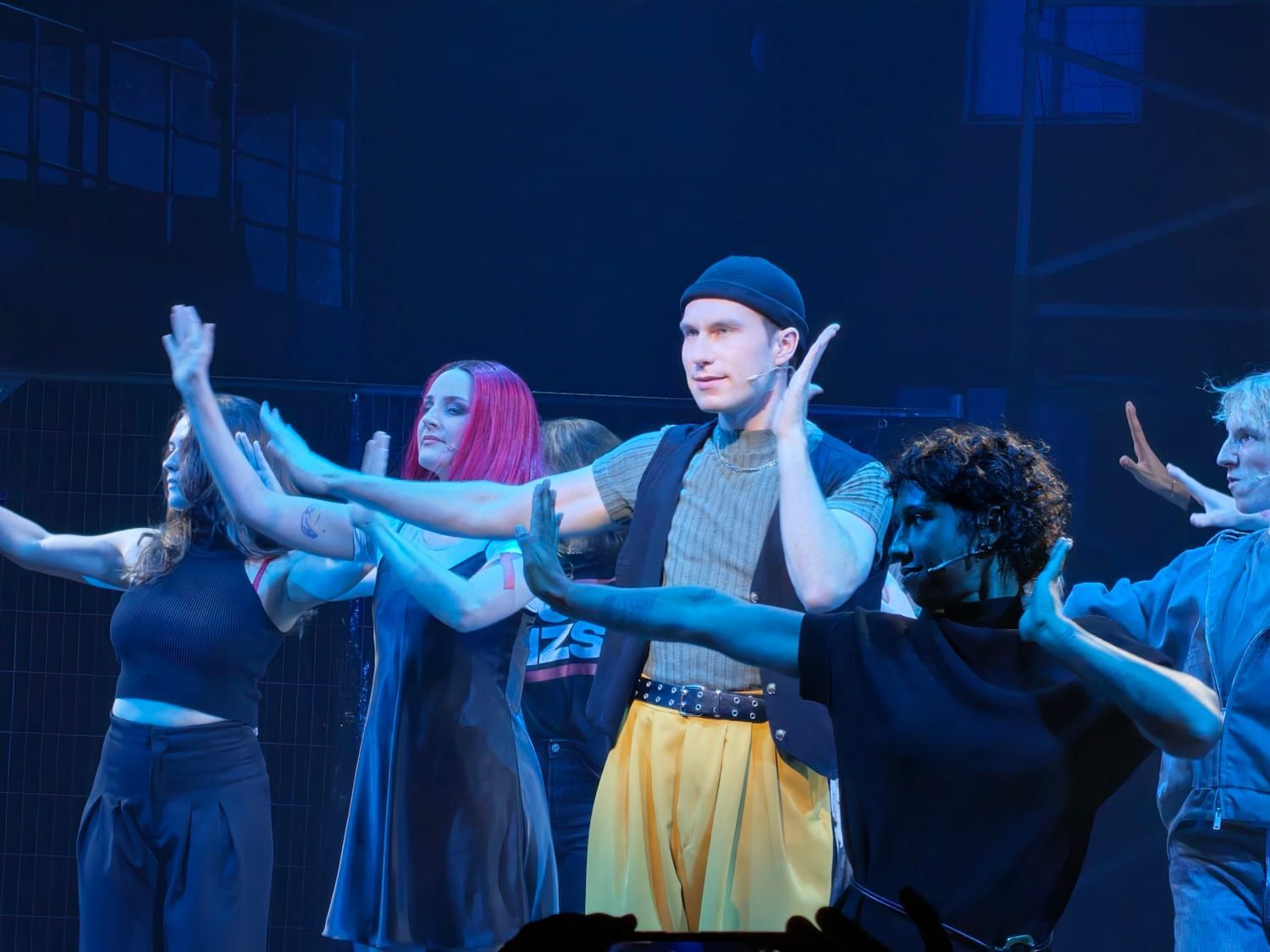 Dancers in blue stage lighting perform synchronized arm movements on a dark theater stage