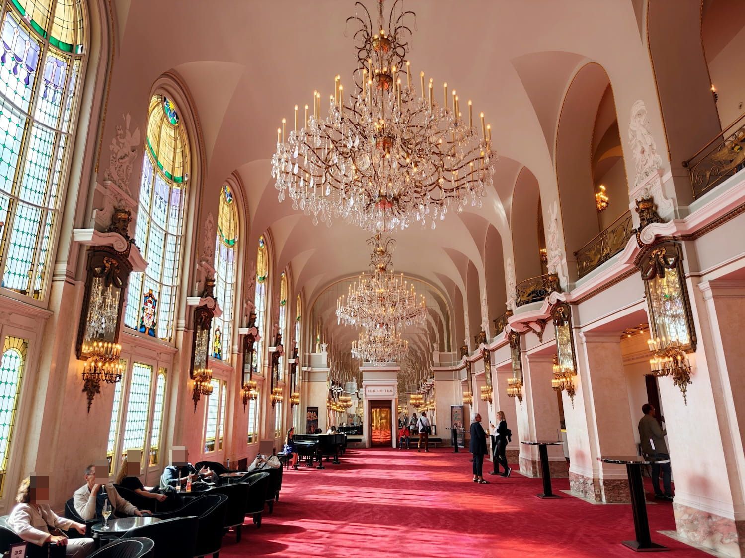 Theater des Westens Grand ballroom with chandeliers, stained-glass windows, red carpet, and people seated along the sides