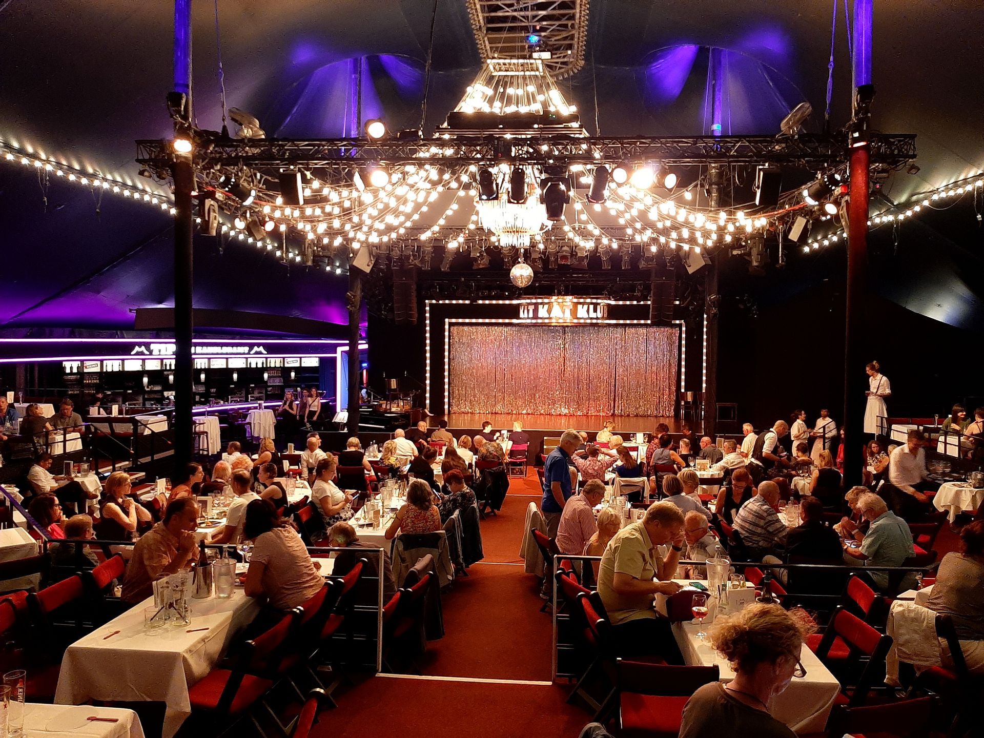 Tipi am Kanzleramt Berlin Crowded banquet hall with long tables, chandeliers, and a brightly lit stage at the far end