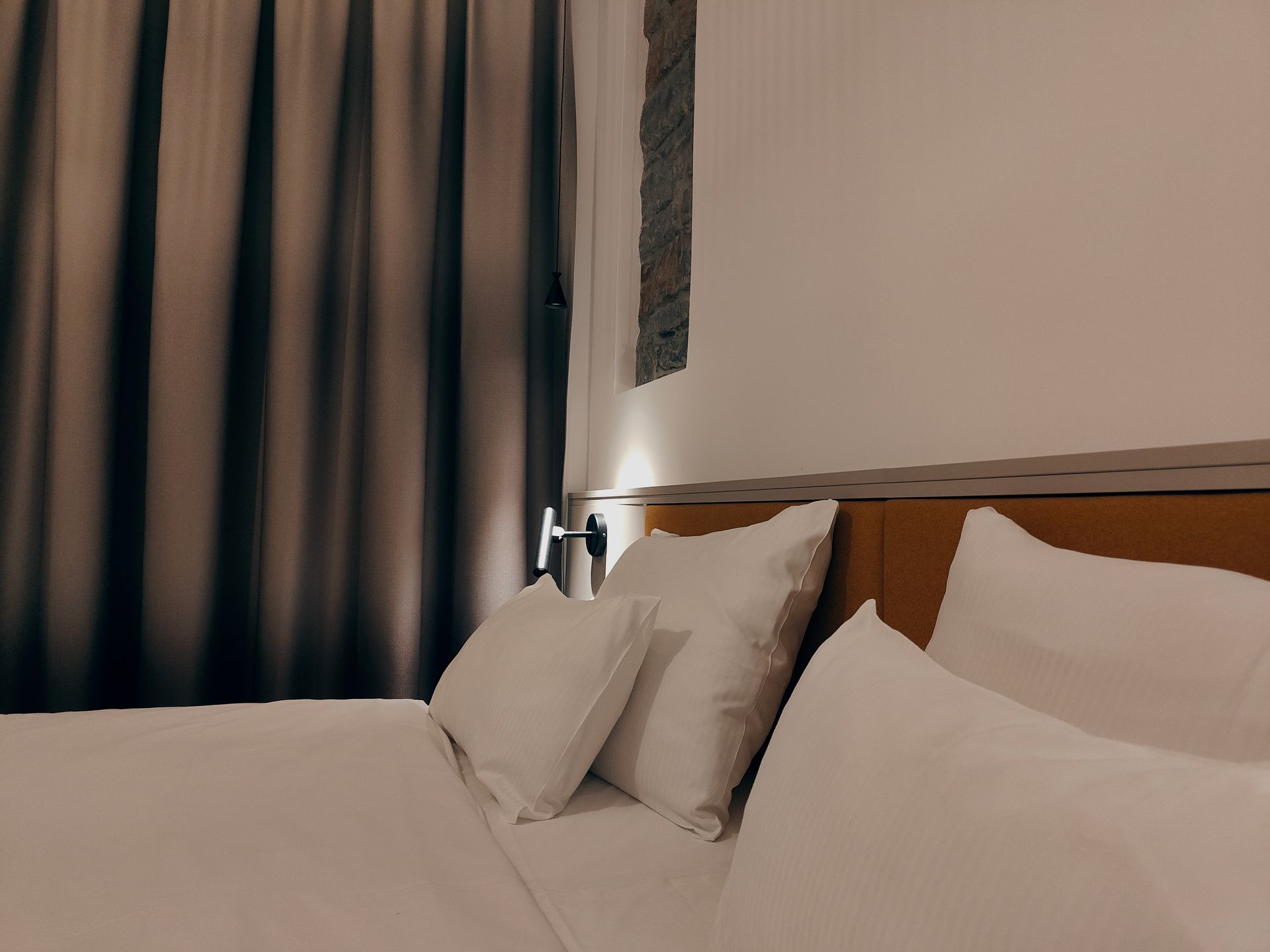 Neatly made bed with white pillows beside dark curtains and a small wall lamp