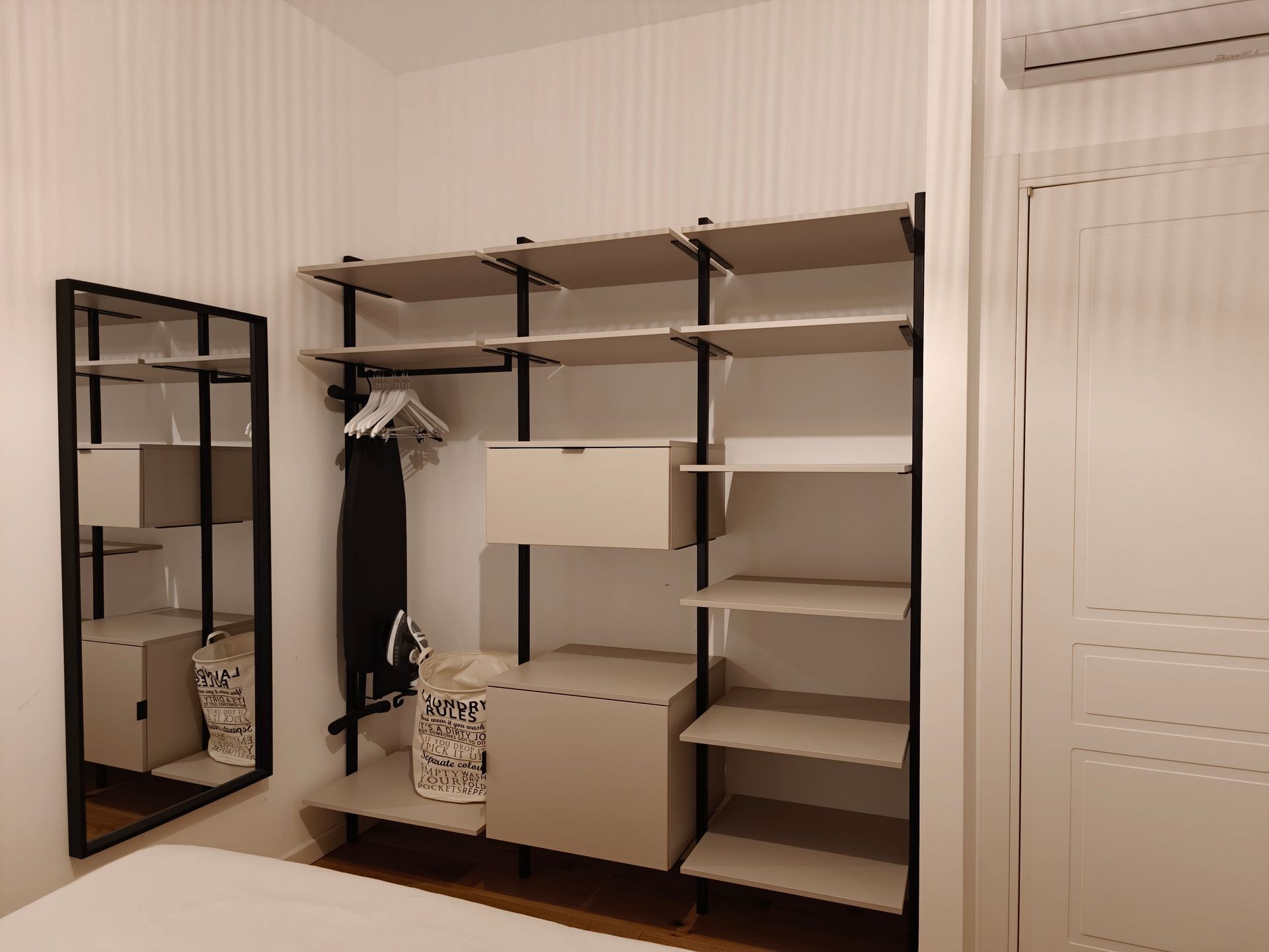 Open closet with white shelves, storage bins, hanging clothes, and a tall mirror beside it.