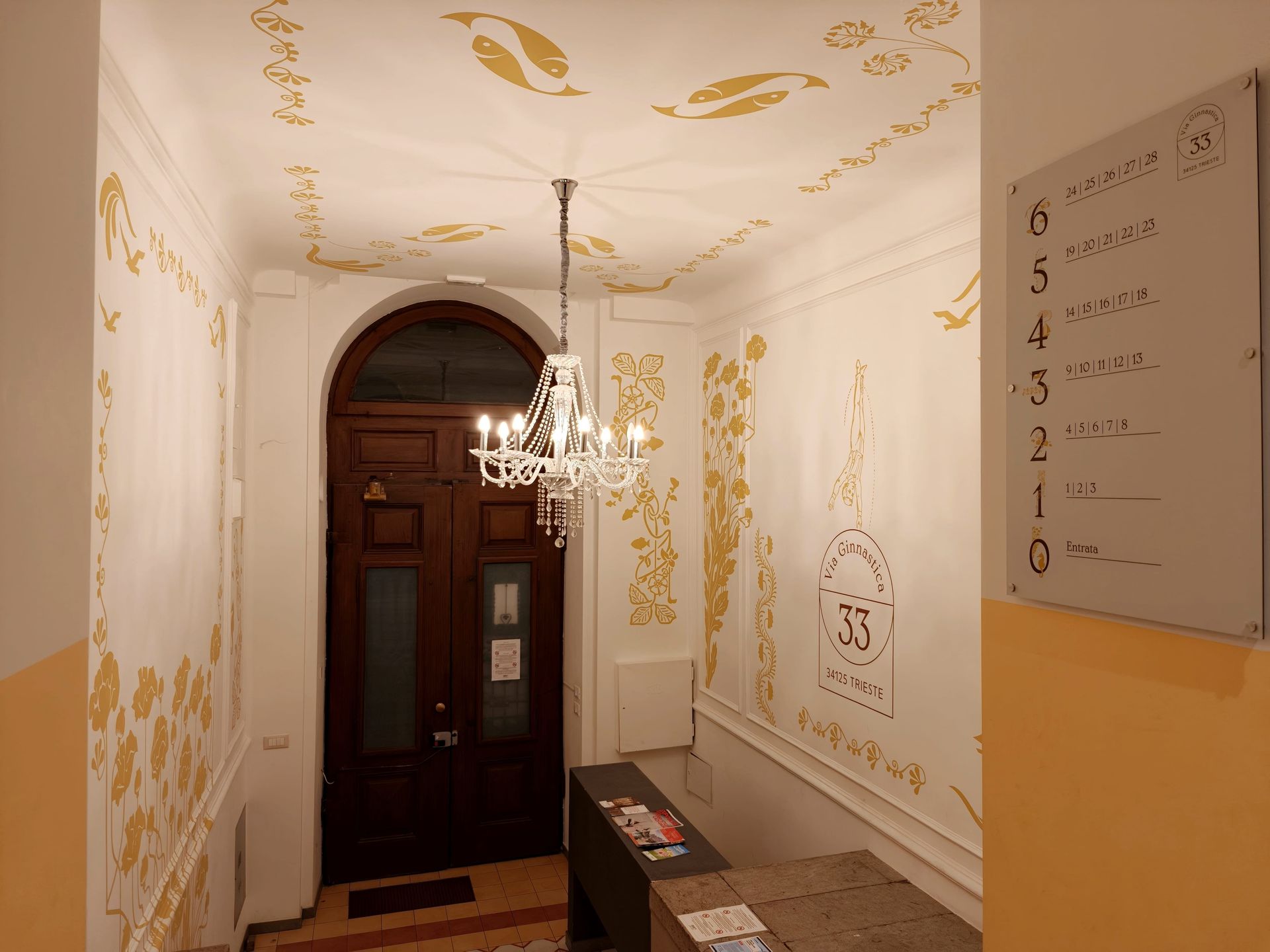 Urbanauts Studios Ginnastica Triest Eingangstor White gallery hallway with gold wall patterns, chandelier, benches, and a dark wooden door at the end