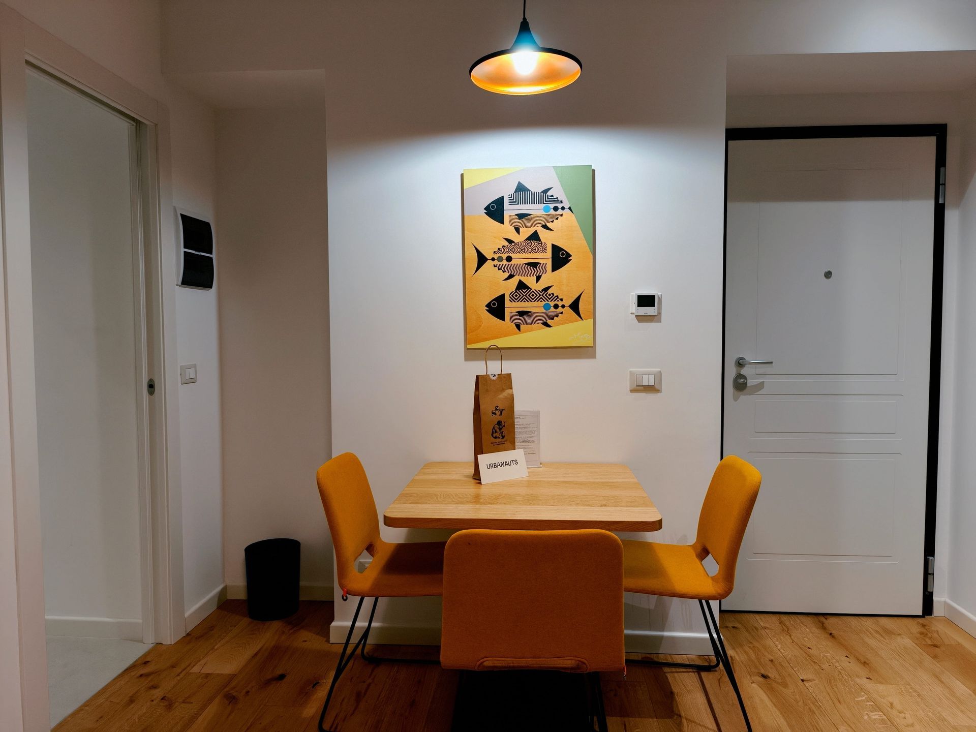 Urbanauts Studios Ginnastica Triest Small dining area with a wooden table, orange chairs, wall art, and white doors under a hanging light.