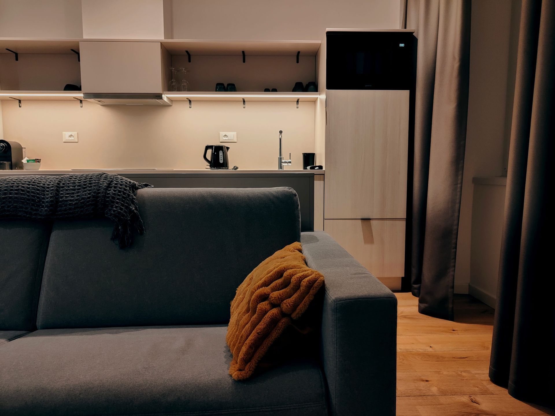 Urbanauts Studios Ginnastica Triest Modern living room with gray sofa, orange cushion, and open kitchen in the background.
