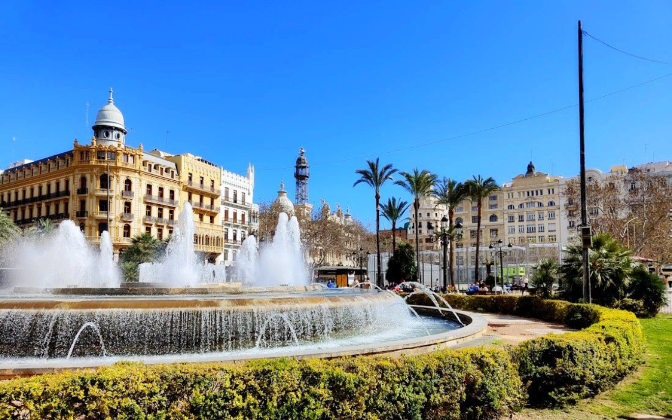 Valencia Fountain in a plaza with buildings and palm trees under a bright blue sky.
