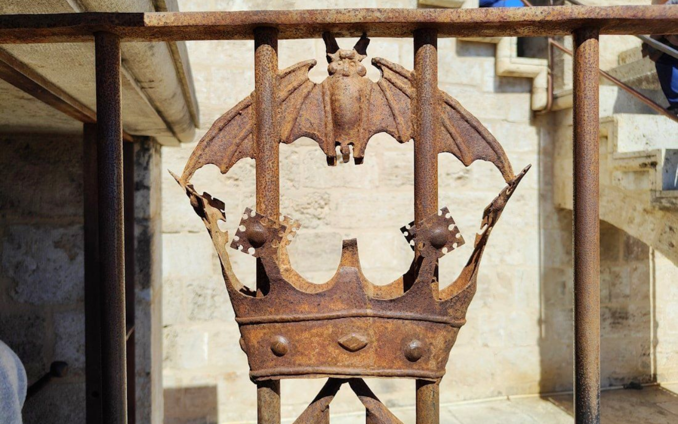 Wappenzeichen Valencias Rusted metal railing detail: a crown with a bat in the center, set against a stone wall and stairs.
