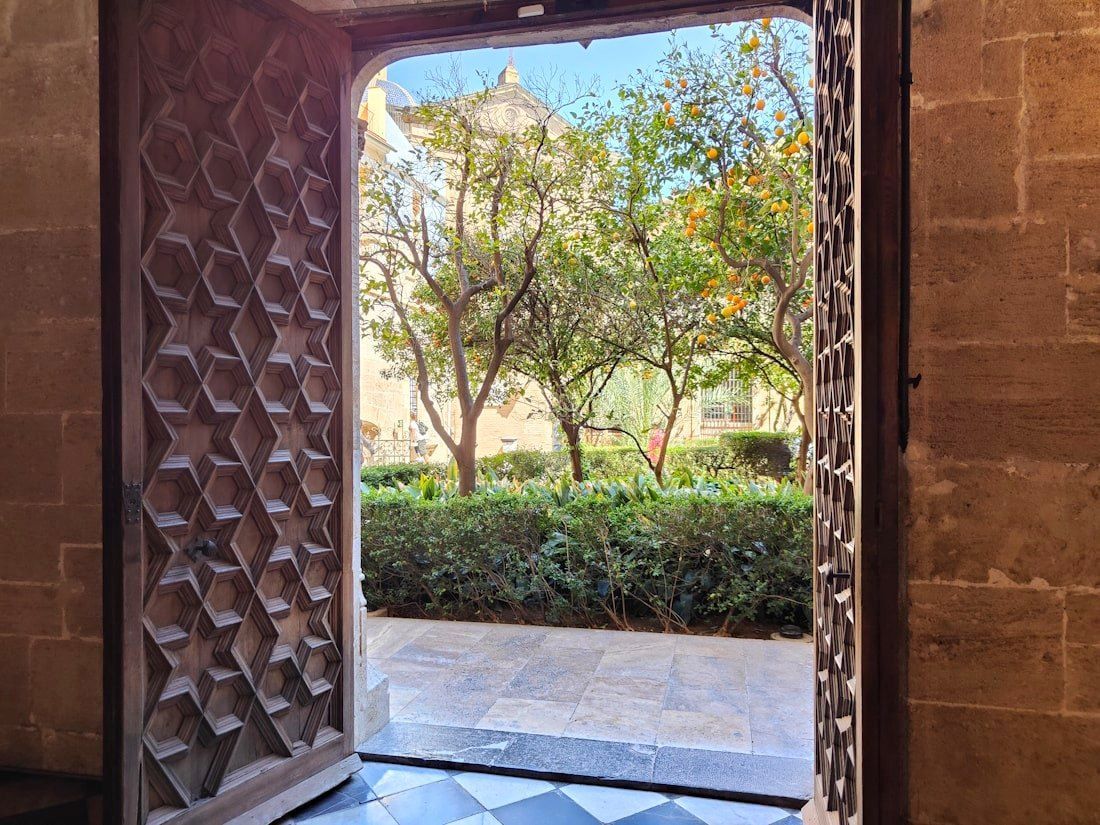 Die Seidenhalle Open wooden doors framing a view of a courtyard with trees, a building, and lush greenery.