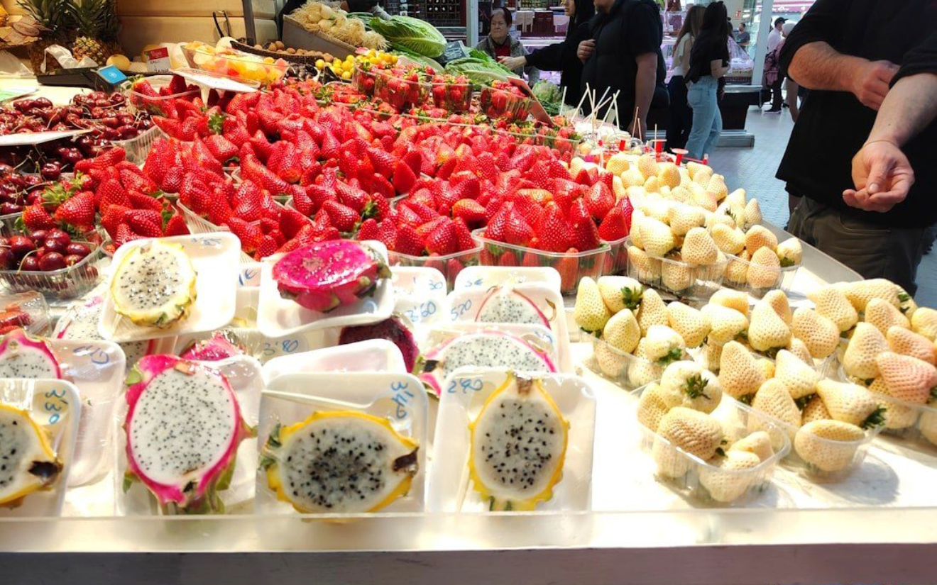 Markthalle Fruit stand display of dragon fruit, white strawberries, and red strawberries. Shoppers browse.