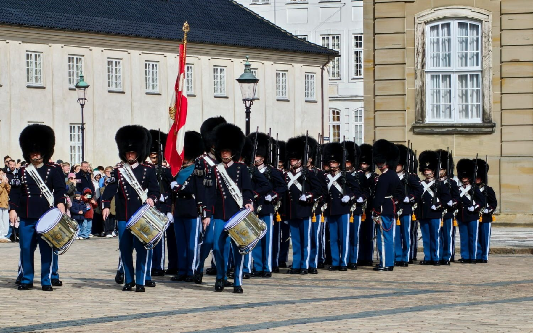 Wachablöse Königliche Garde Royal guards in blue uniforms and bearskin hats march with drums in a courtyard, carrying a red flag. Wachablöse Königliche Garde Kopenhagen