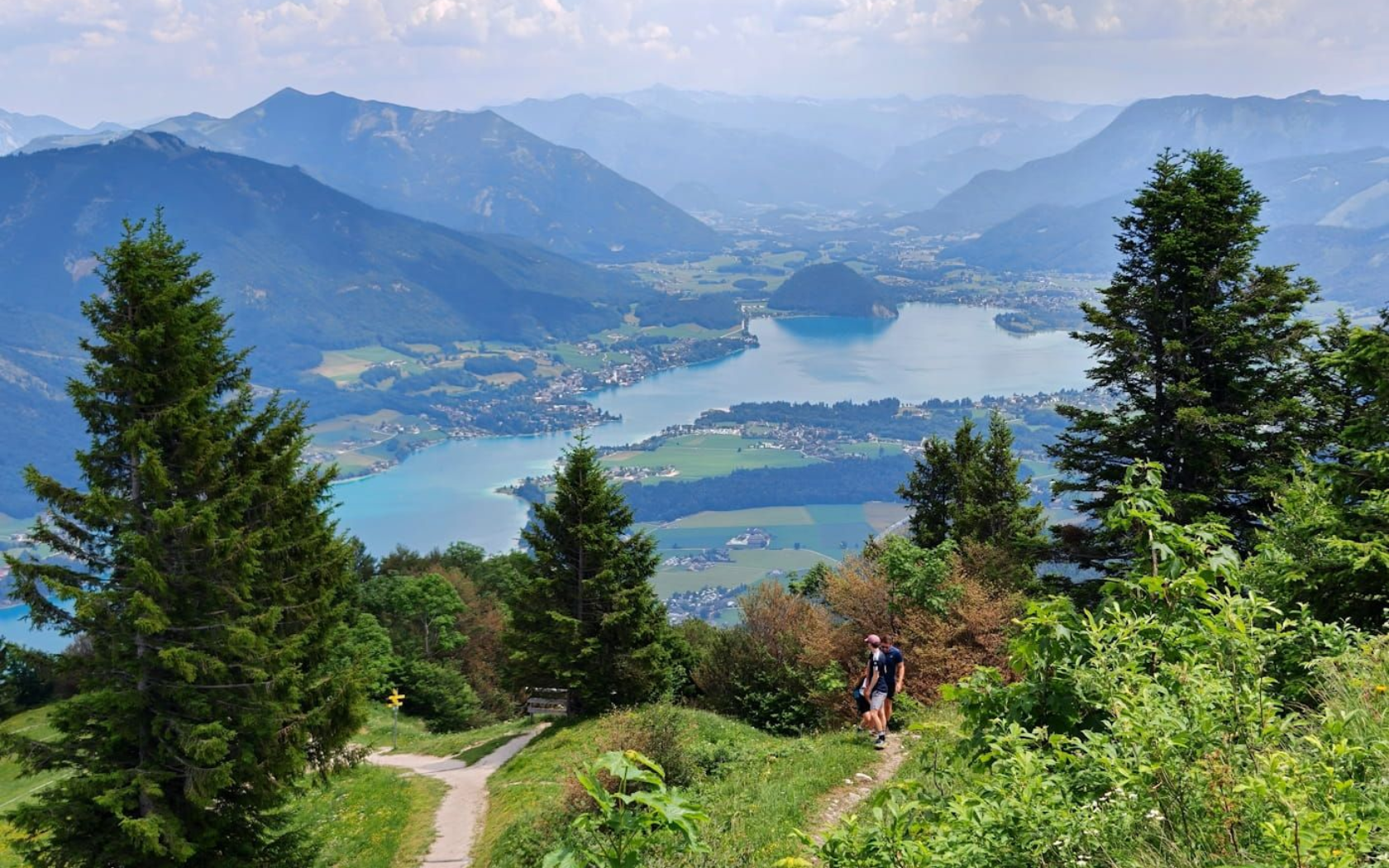 Wolfgangsee Mountain trail overlooking a blue lake, valley, and distant Alps under a hazy sky