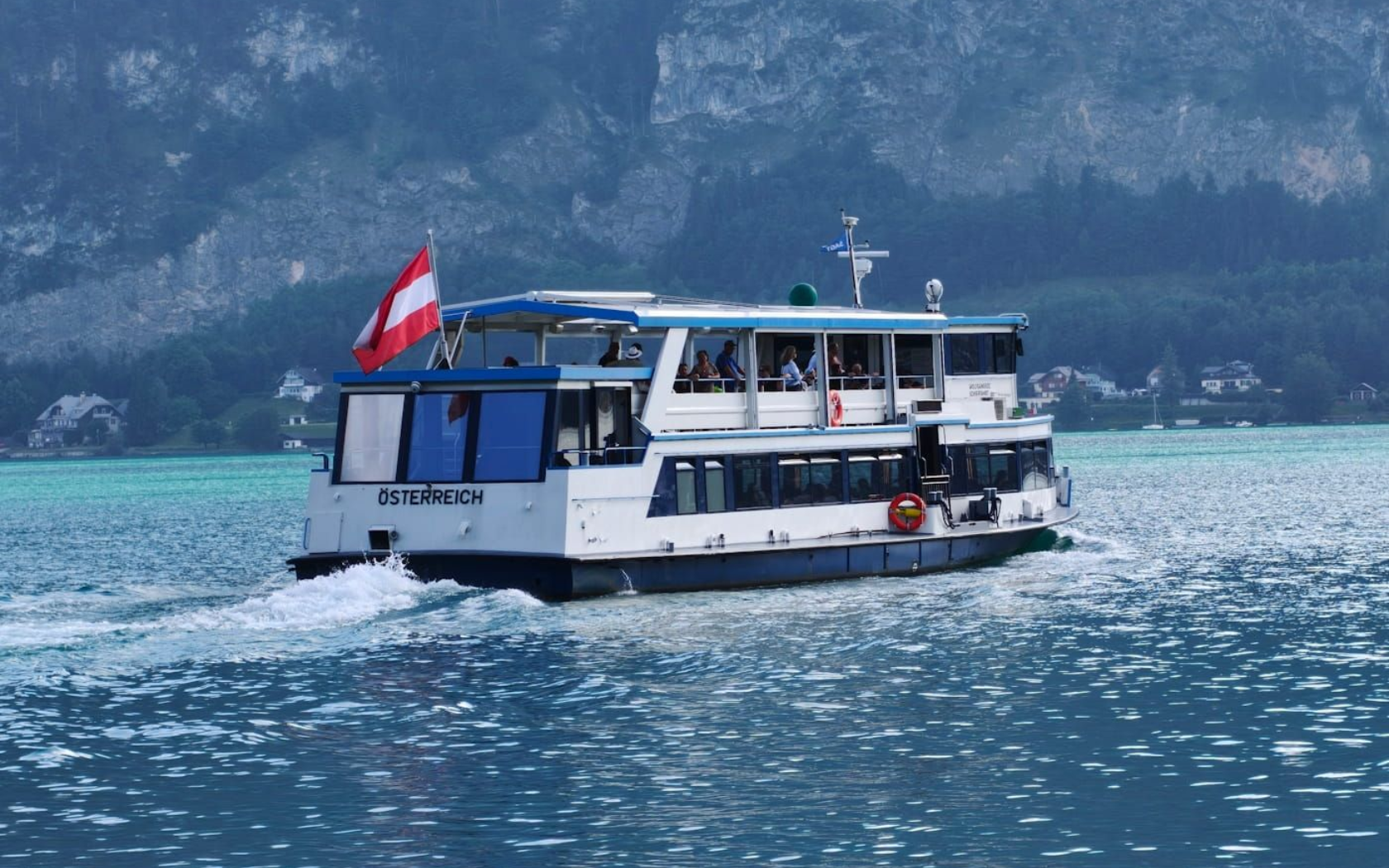 WolfgangseeSchifffahrt White ferry boat with a red flag sails on turquoise water in front of a forested shoreline