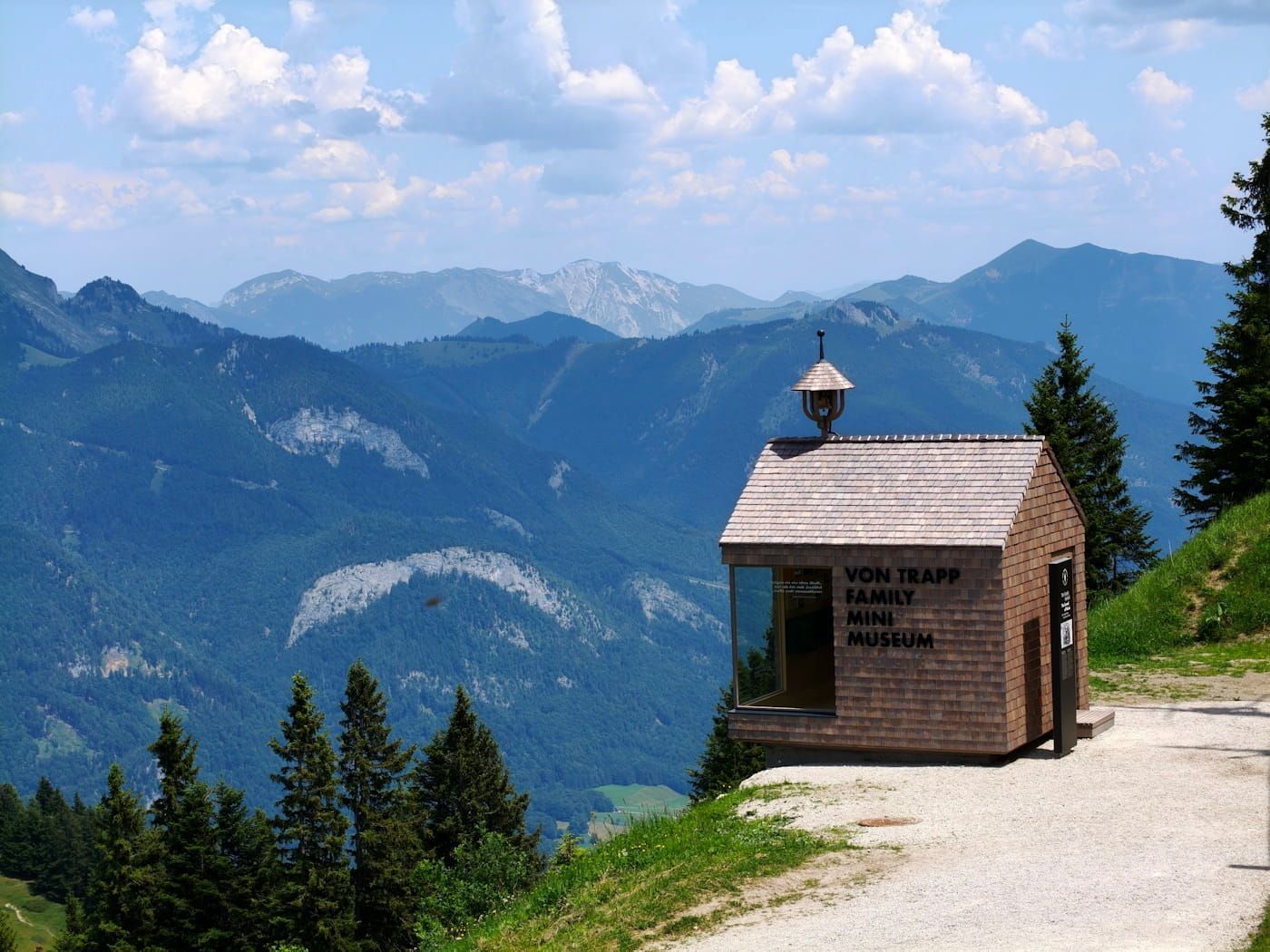 Small wooden chapel on a mountain trail with a scenic view of forested peaks and blue sky.
