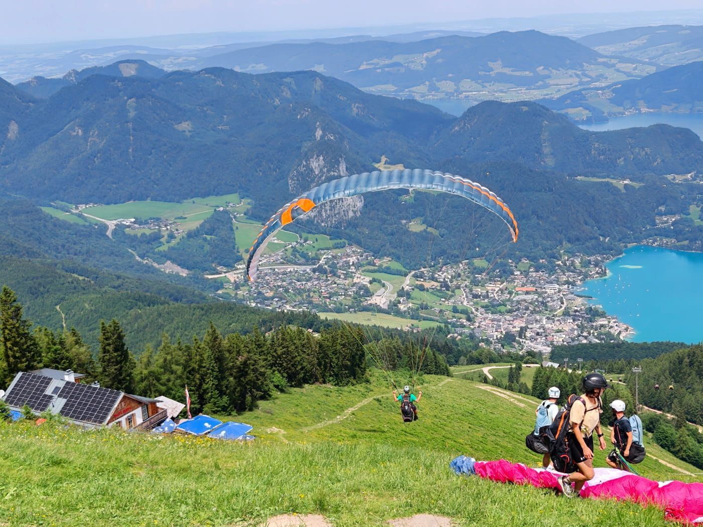 Paragliders over a mountain town and blue bay, viewed from a grassy hillside with people resting.