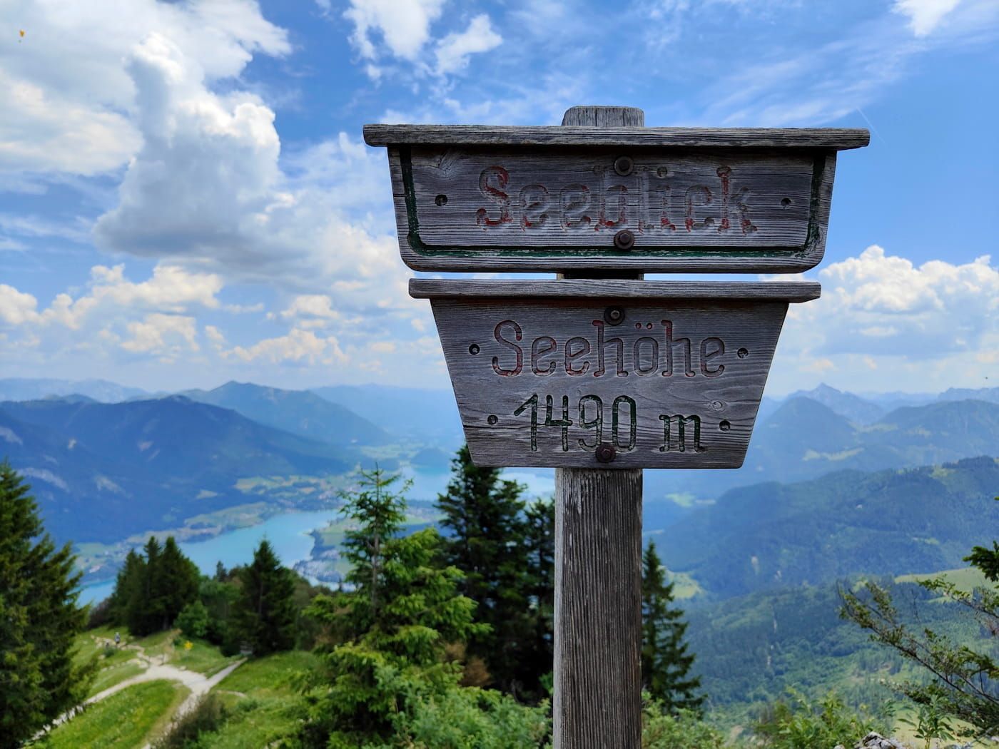 Wooden trail sign overlooking a lake and mountains under a cloudy blue sky