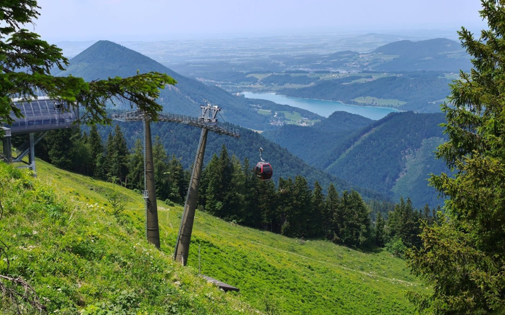Zwölferhorn Seilbahn Green hillside with ski lift poles overlooking a distant valley and mountains under a hazy blue sky