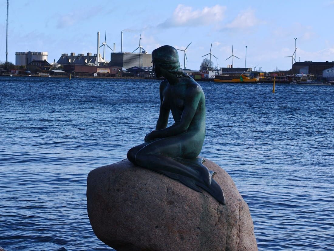 Bronze mermaid statue on a rock by the water with an industrial shoreline in the background