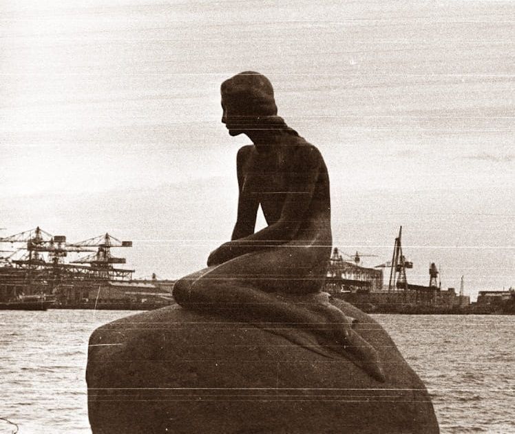 Silhouette of a seated statue by the water with cranes and ships in the background