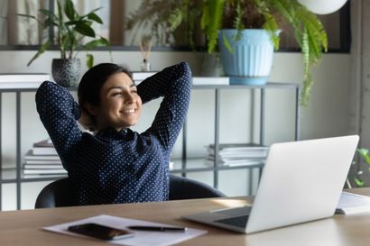 A woman is sitting at a desk with her hands behind her head in front of a laptop computer.