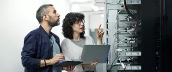 A man and a woman are looking at a laptop in a server room.