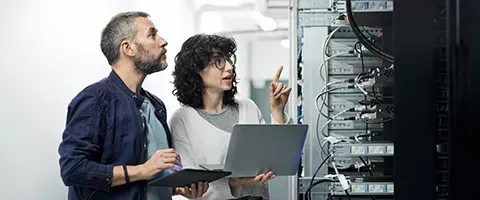A man and a woman are looking at a laptop in a server room.