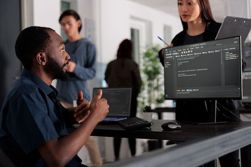 A man is sitting at a desk in front of a computer talking to a woman.