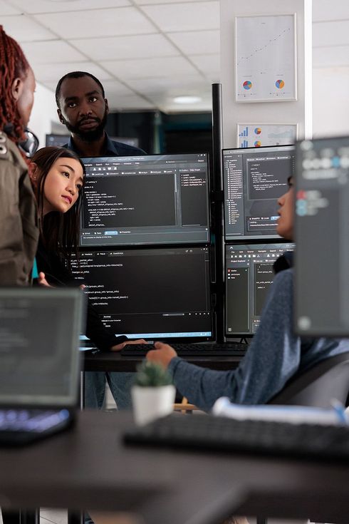 A group of people are standing around a desk in front of computer monitors.