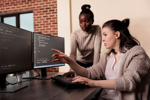 A woman is pointing at a computer screen while another woman looks on.