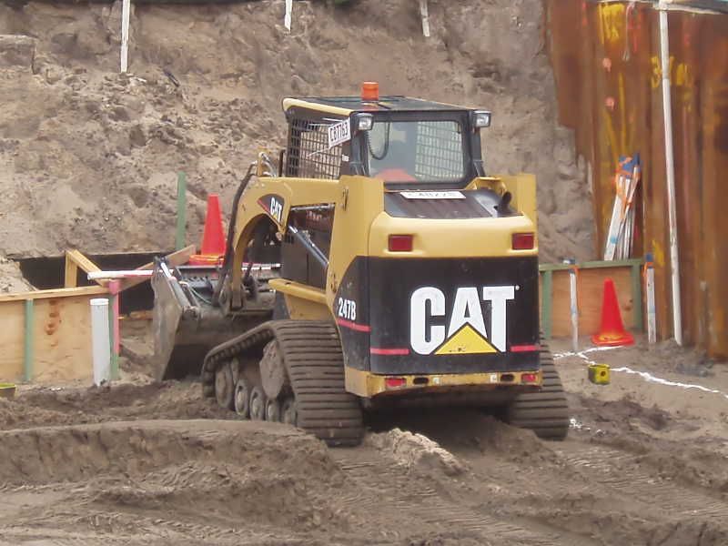 Yellow CAT excavator digging in a muddy construction trench, with traffic cones nearby