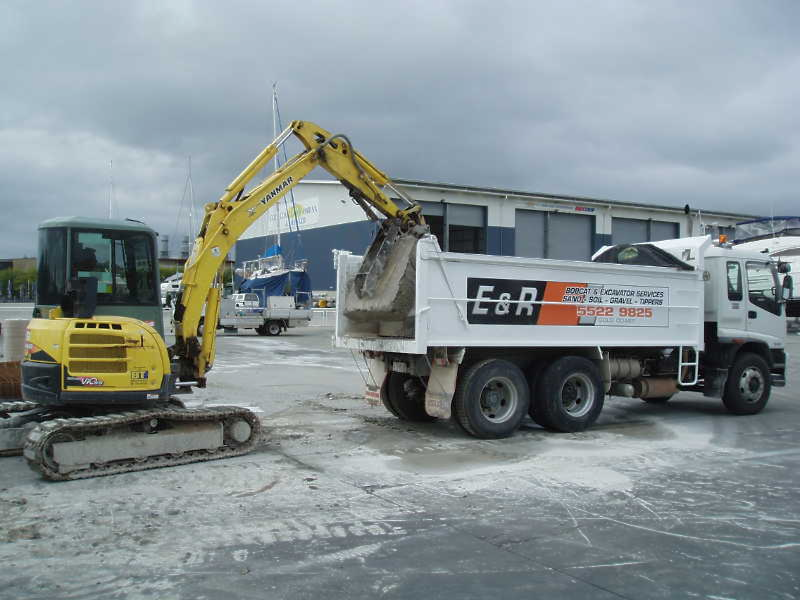 Yellow excavator loading soil into a white dump truck at a snowy construction site