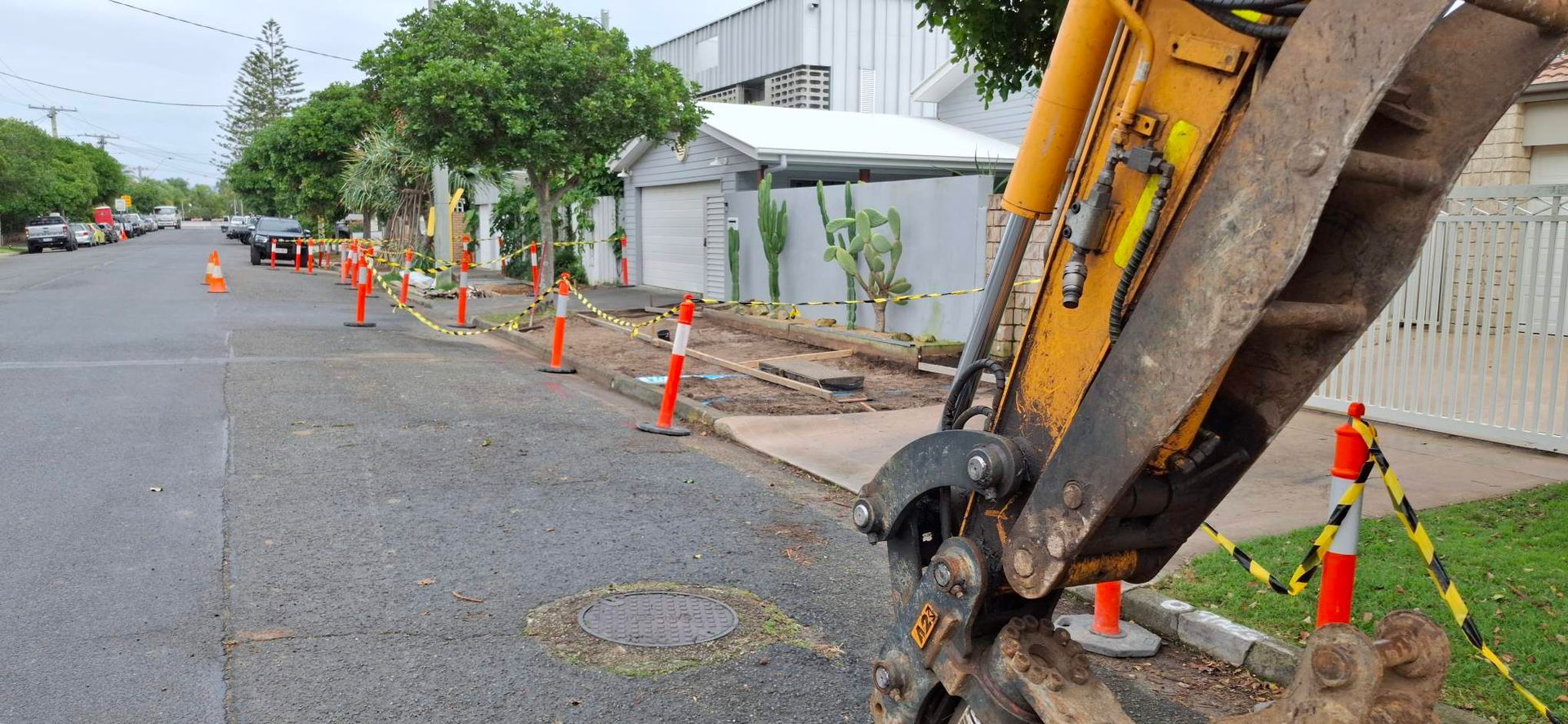 Excavator working on roadside with orange cones beside a sidewalk and apartment building.