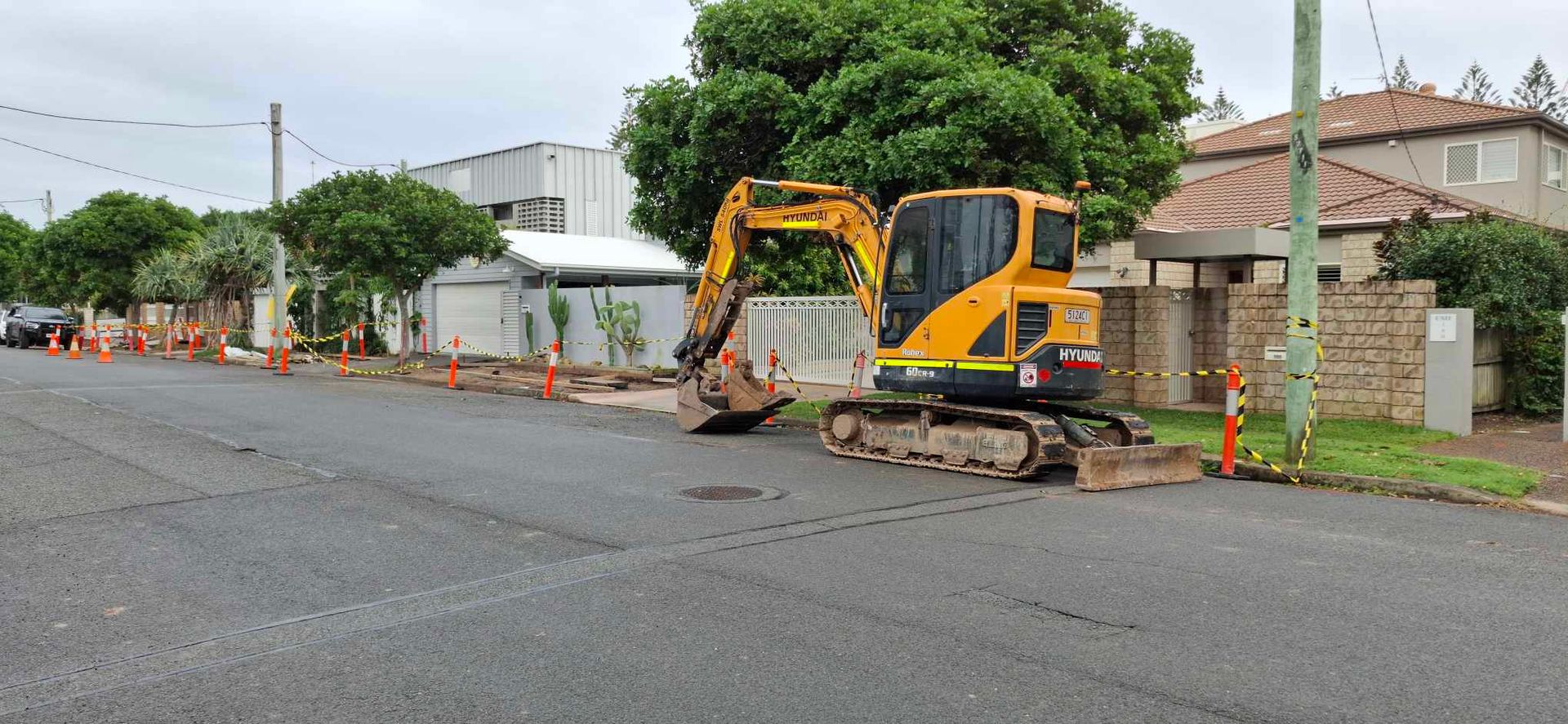 Yellow excavator parked on a wet residential street beside trees and houses
