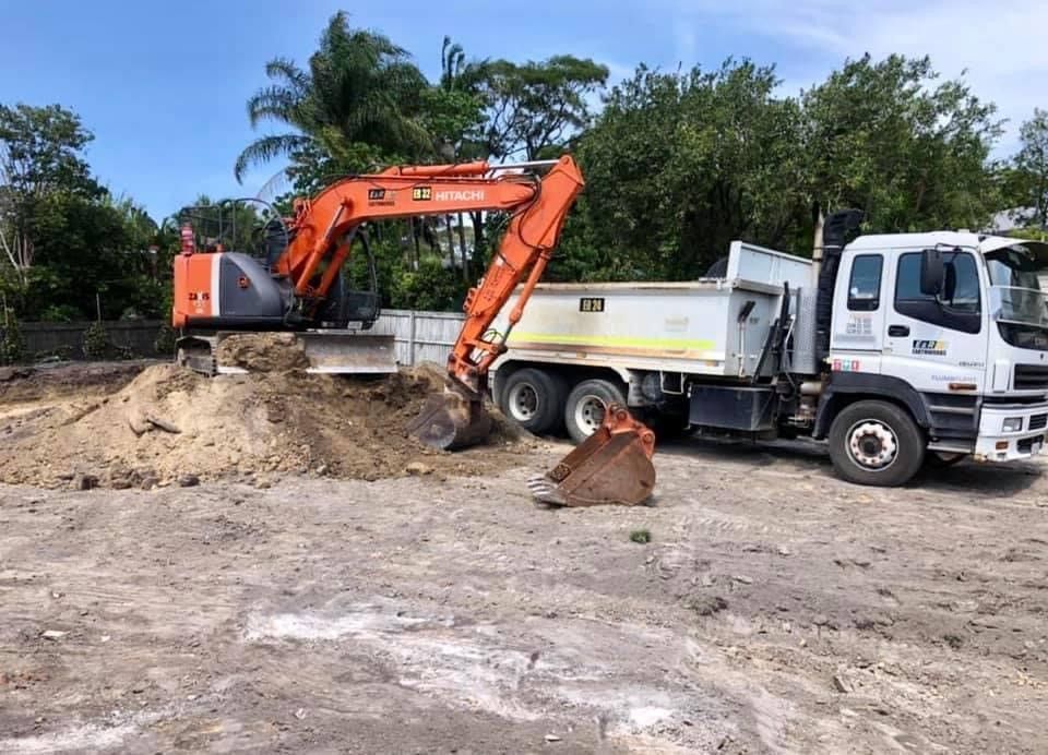 Orange excavator loading dirt into a white dump truck at a construction site, with trees in the background