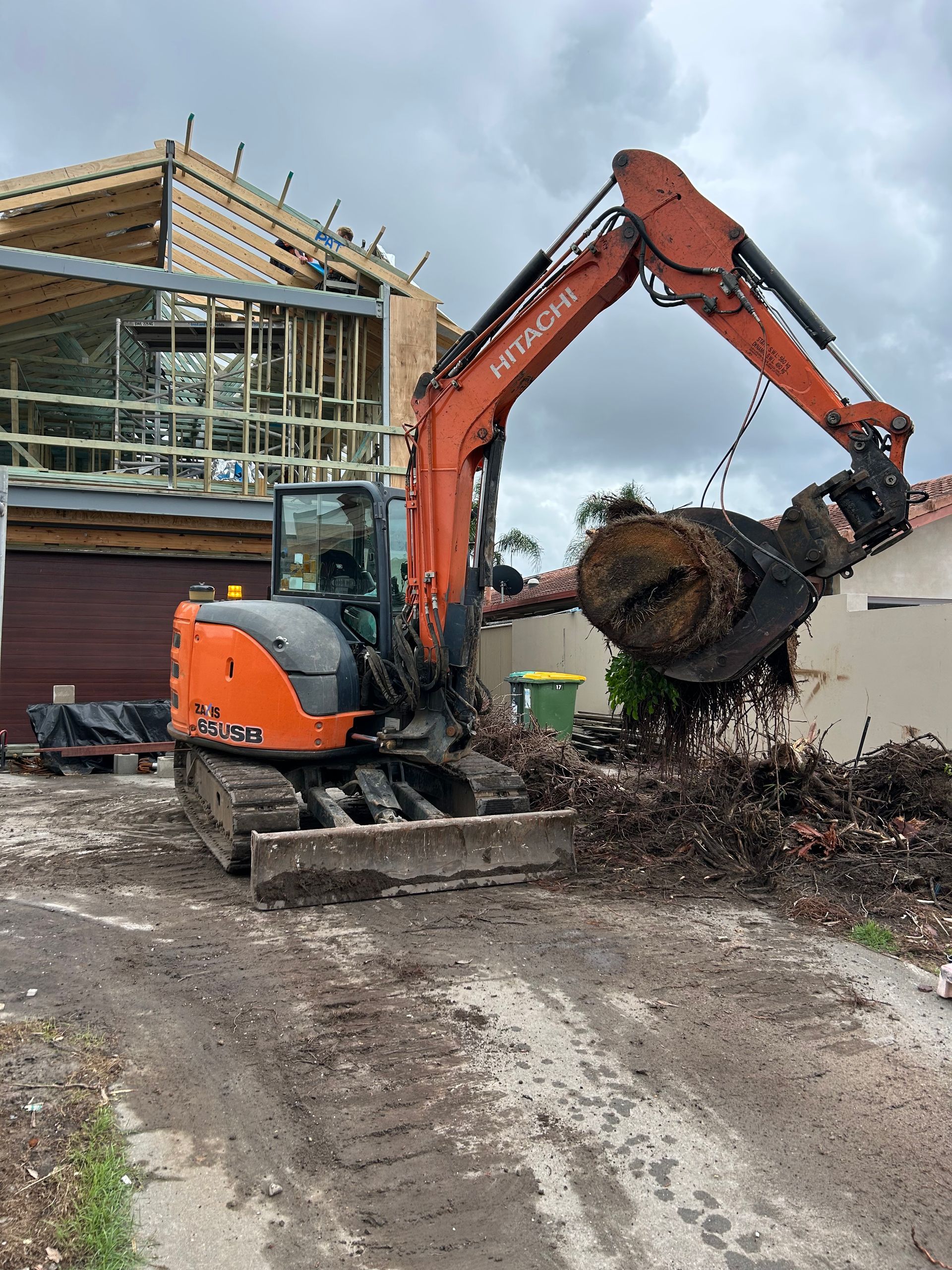 Orange excavator lifting a large tree stump at a muddy construction site beside a house under construction