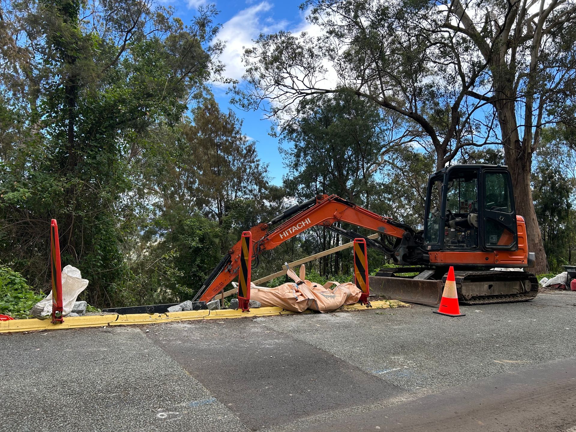 Small orange excavator and cone on a gravel roadside beside a trench, with trees in the background.