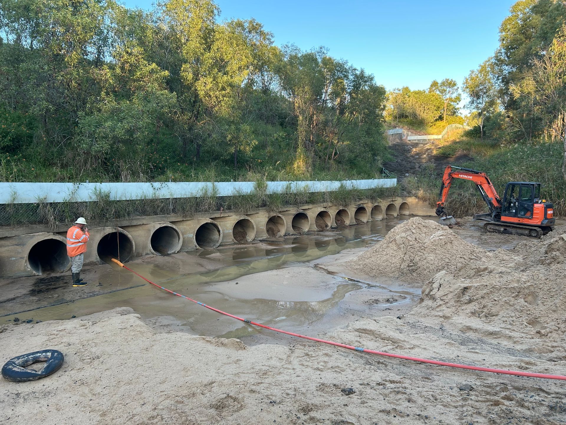 Construction site with orange excavator and worker beside large drainage culvert pipes along a dirt embankment