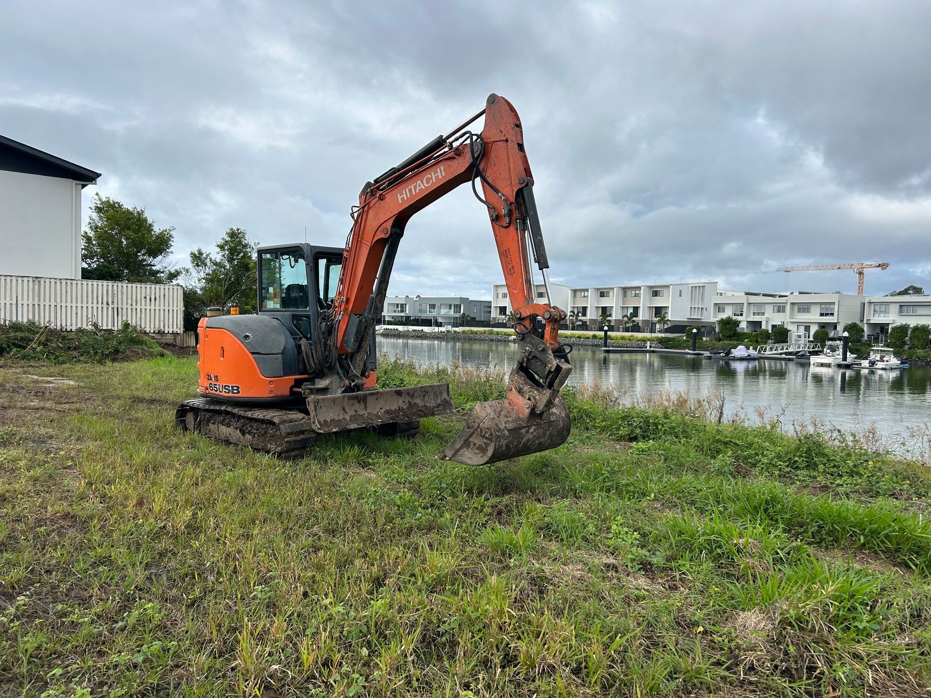 Orange excavator clearing wet grass beside a pond under a cloudy sky